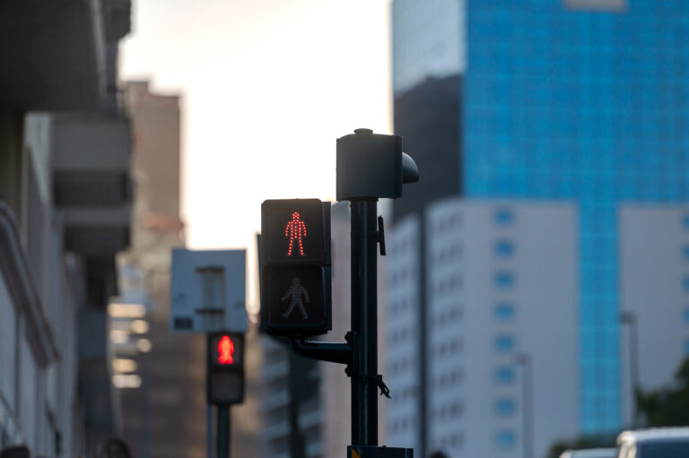 A red pedestrian signal indicates stop while urban buildings rise in the background.